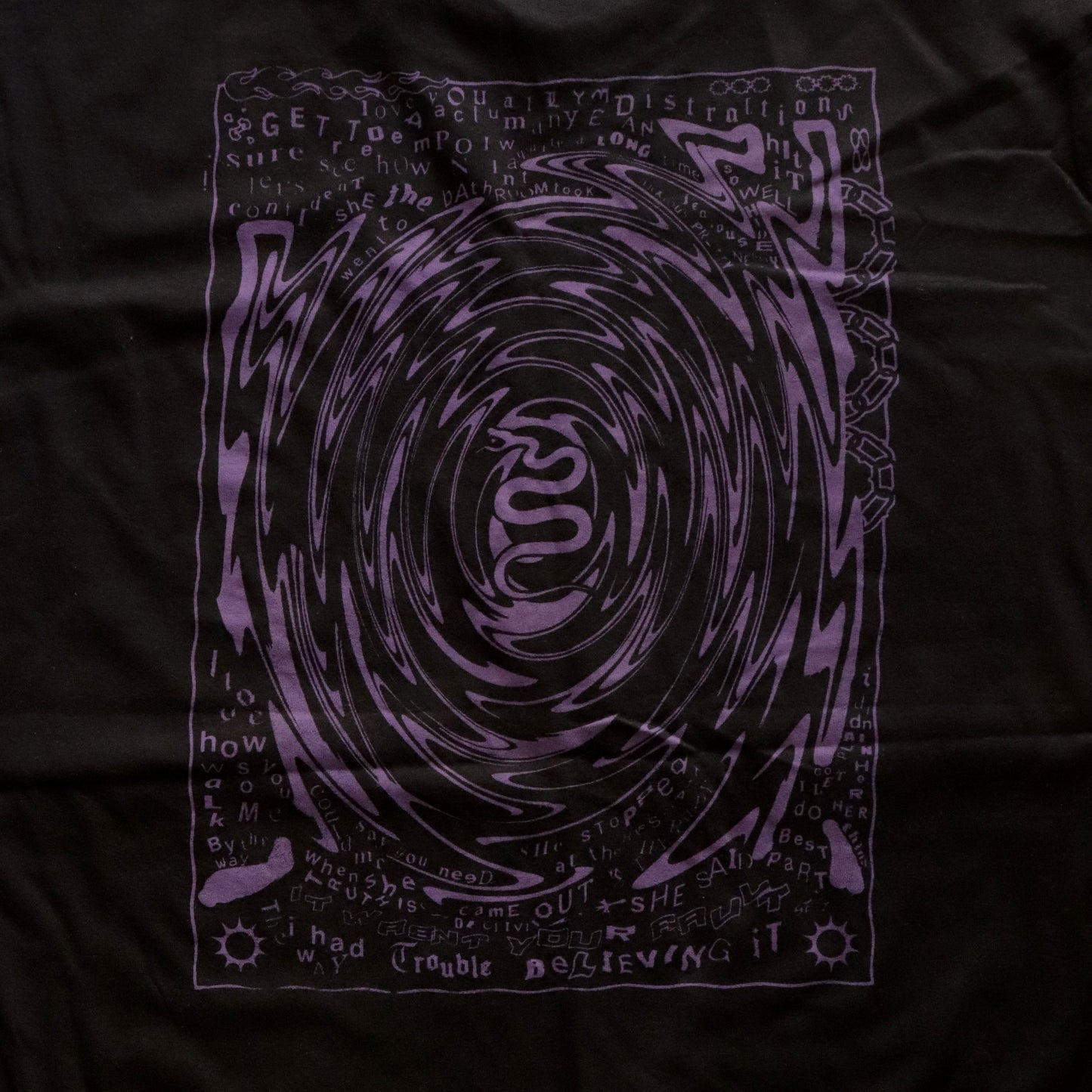 Close-up of Black t-shirt featuring a purple hand screen-printed design with swirling artwork and handwritten lyrics from the song ‘FILTH’ on the back.”