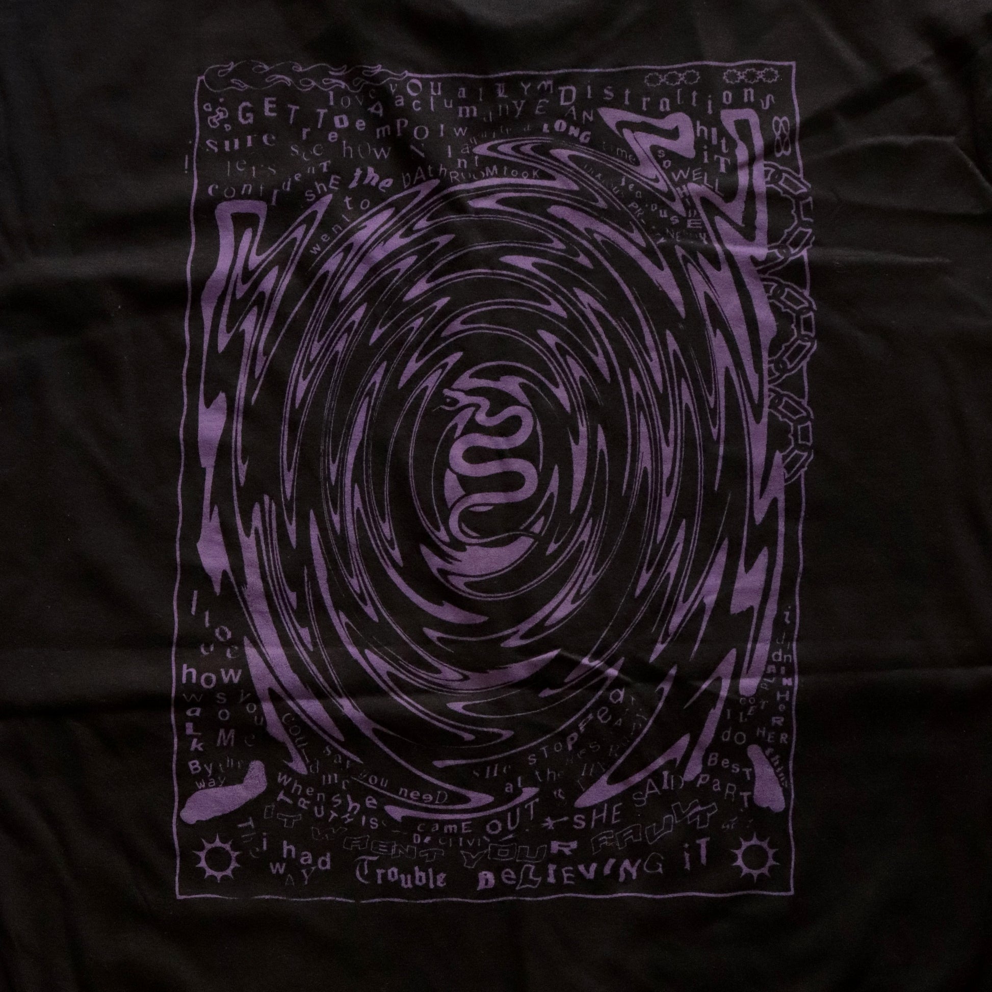 Close-up of Black t-shirt featuring a purple hand screen-printed design with swirling artwork and handwritten lyrics from the song ‘FILTH’ on the back.”
