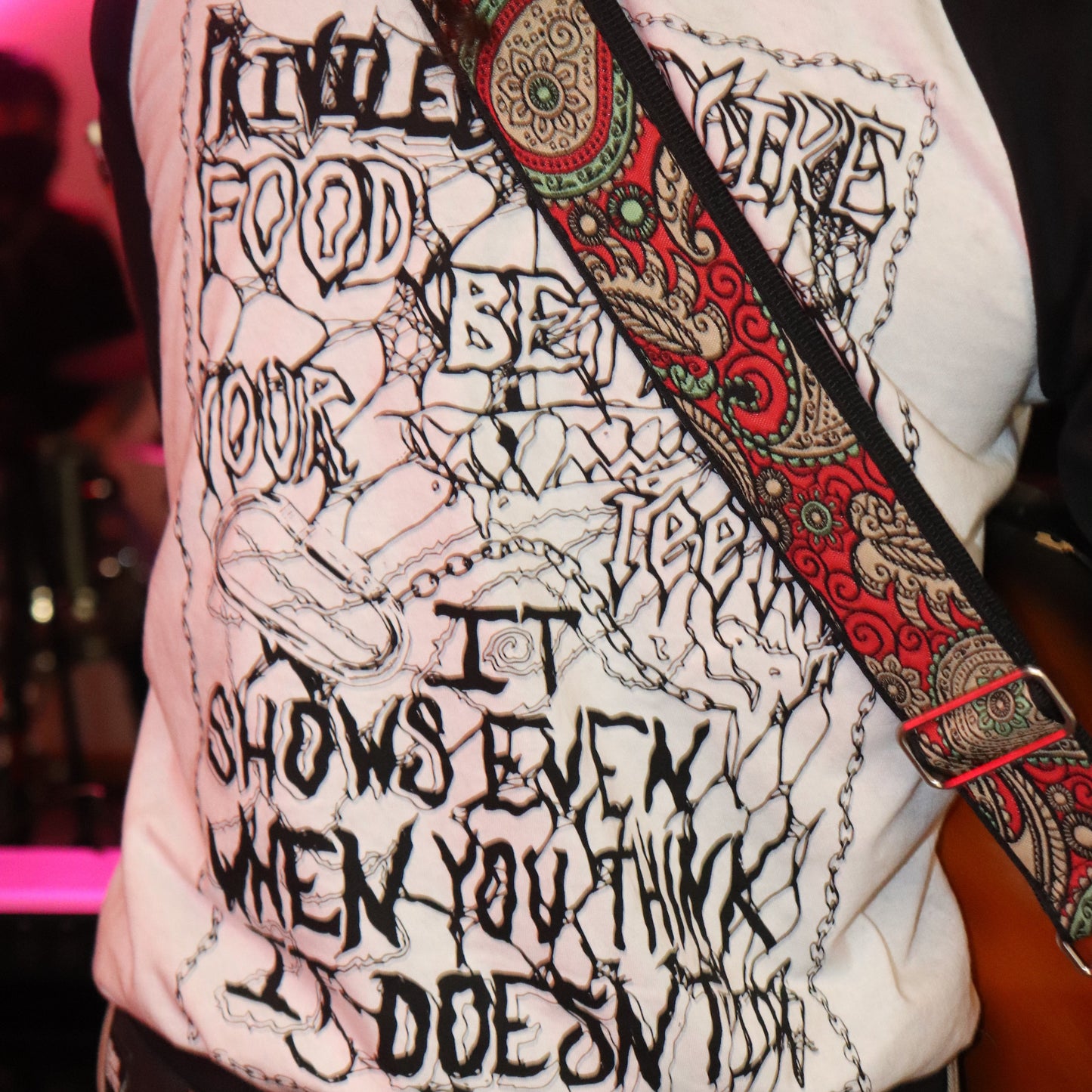 Person wearing a white shirt with black text and graphics, holding a guitar with a colorful strap.