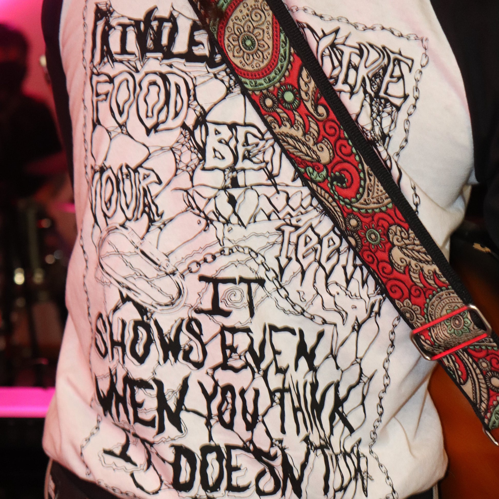 Person wearing a white shirt with black text and graphics, holding a guitar with a colorful strap.